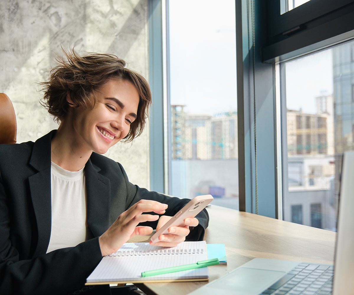 Portrait of young professional saleswoman, businesswoman in suit, sitting in office at her company, using smartphone, working on laptop.